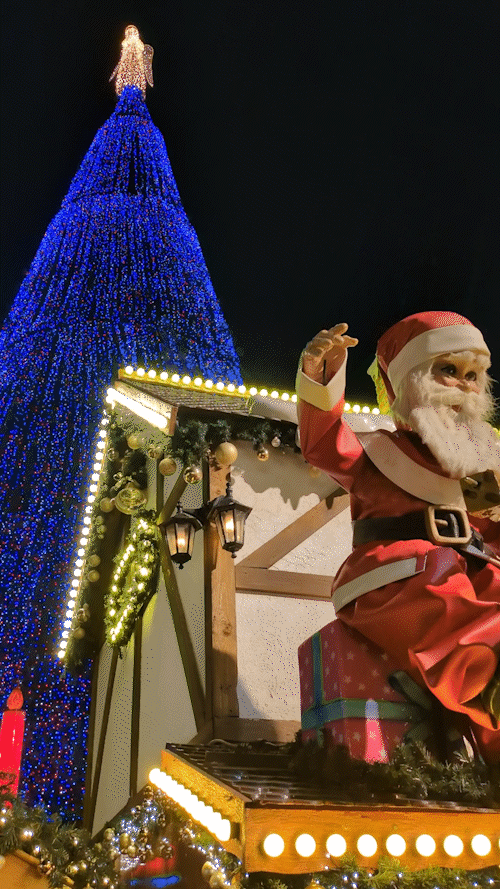 A gif of a robot Santa holding a stick full of cheese cubes in front of the world's largest Christmas tree